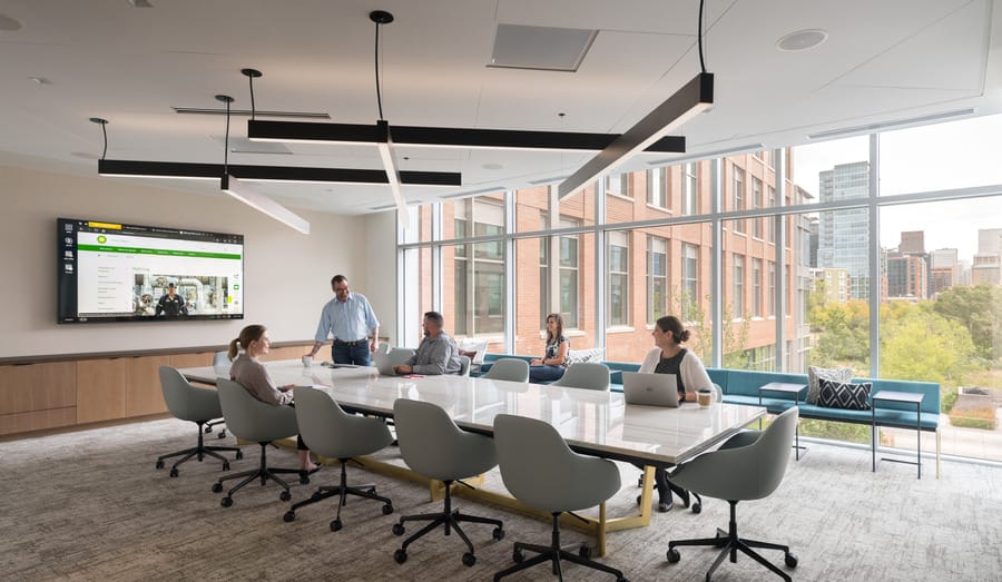 A conference room with a group of people having a meeting with a television displaying content on the wall. A wall of windows is behind them.