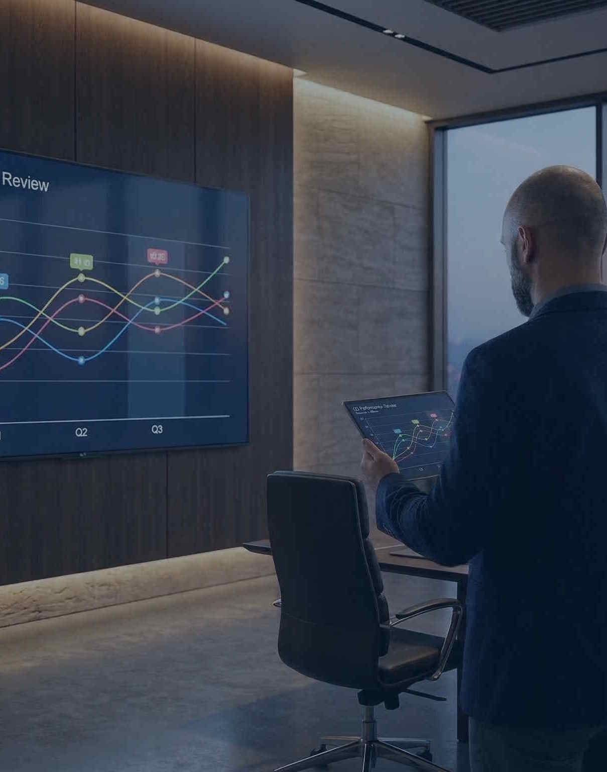 Businessman reviewing performance data on a tablet and large display in a conference room